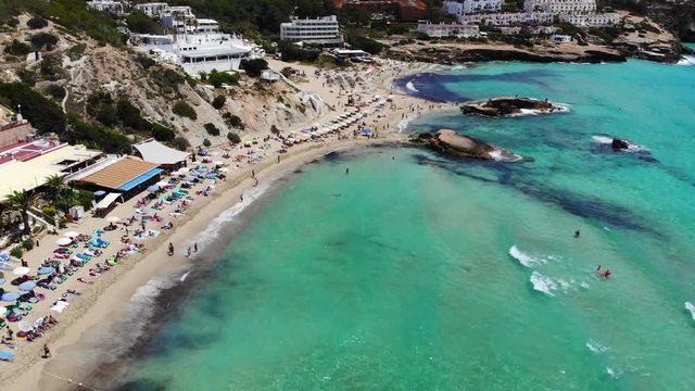 drone shot tilting down at cala tarida beach in ibiza with people at the beach sunbathing and swimming