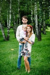 Fototapeta premium Mother, Dad hold a son on the arms in embroidery. Happy family on the background of birches on nature. Close up. Looking at the camera. Full length. Embroidered shirt and handmade dress.