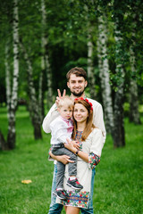 Fototapeta na wymiar Dad Mother hold son on arms in embroidery. Happy family on background of birches on nature. Close up. Looking at the camera. upper half. Embroidered shirt and handmade dress. putting horns on head.