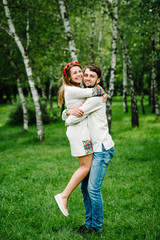 The man raised the woman to her arms.  Happy family on the background of birches on nature. Close up. Looking at the camera. Full length. Embroidered shirt and handmade dress.