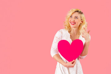 Studio portrait of a laughing young woman holding pink heart, looking and pointing up at copy space, Valentine's Day love concept