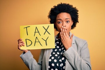 Young African American afro woman with curly hair holding paper with tax day message cover mouth...