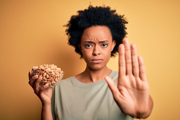 Young African American afro woman with curly hair holding bowl with salty peanuts with open hand...