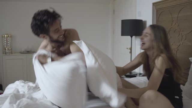Young attractive couple having a silly fun pillow fight on bed, laughing and loving.
