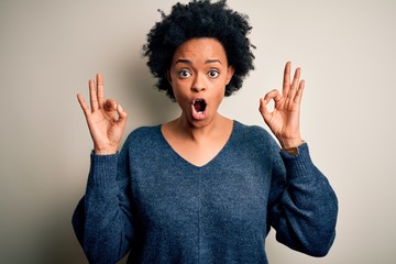 Young beautiful African American afro woman with curly hair wearing casual sweater looking surprised and shocked doing ok approval symbol with fingers. Crazy expression