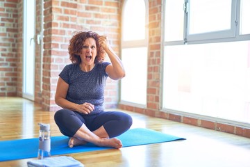 Middle age beautiful sportswoman wearing sportswear sitting on mat practicing yoga at home angry and mad raising fist frustrated and furious while shouting with anger. Rage and aggressive concept.
