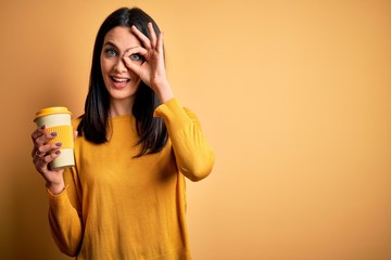 Young woman with blue eyes holding cup of coffee standing over yellow background with happy face smiling doing ok sign with hand on eye looking through fingers