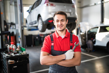 Auto service. Handsome young auto mechanic in uniform is looking at camera and smiling examining car