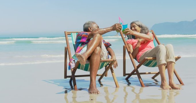 Front view of active senior African American couple toasting drinks on deckchair on the beach 4k