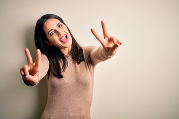 Young brunette woman with blue eyes wearing casual sweater over isolated white background smiling with tongue out showing fingers of both hands doing victory sign. Number two.