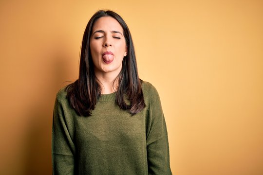 Young brunette woman with blue eyes wearing green casual sweater over yellow background sticking tongue out happy with funny expression. Emotion concept.