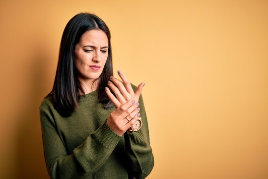 Young brunette woman with blue eyes wearing green casual sweater over yellow background Suffering pain on hands and fingers, arthritis inflammation