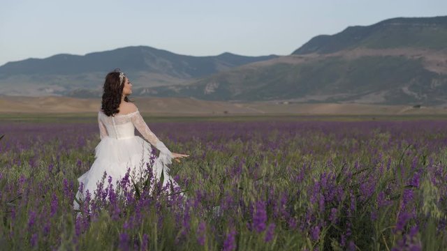 attractive young lady in stylish wedding dress poses among wild lavender purple flowers against mountains slow motion