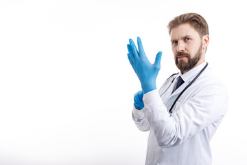 Confident bearded physician in white smock putting on blue medical gloves isolated white background copyspace