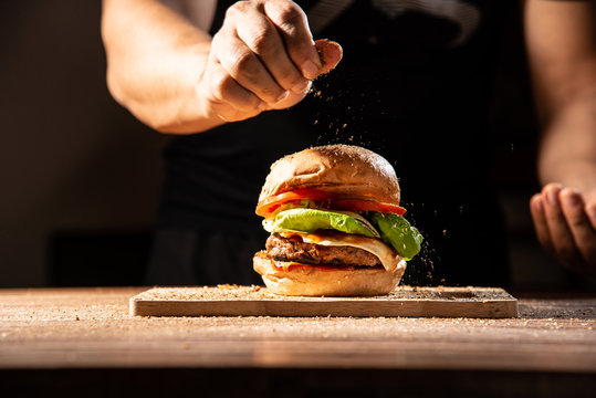 Make it pro, home made hamburger made by white sesame bun, tomato slice, salad, cheese, grilled meat and onion on wooden tray wooden table with cooking decorate chef hand in dark isolate background