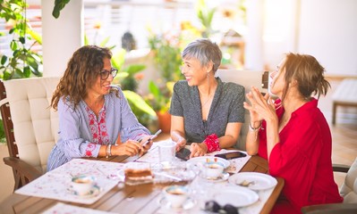 Meeting of middle age women having lunch and drinking coffee. Mature friends smiling happy using smartphone at home on a sunny day