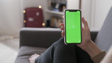 Close up of a female's hand holding a mobile telephone indoors. Young woman sitting on a couch with green screen smartphone in vertical. Browsing Internet watching video. Mock-up for tracking content