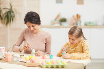 Fototapeta premium Candid portrait of mother and daughter painting Easter eggs pastel colors sitting at table in cozy kitchen interior, copy space
