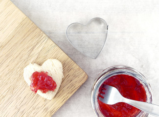 Fototapeta na wymiar strawberry jam spread on small heart shape white bread on cutting board, pastry cutter and berry jam in glass jar with silver fork on blotting paper, wedding appetizer menu, close up top view