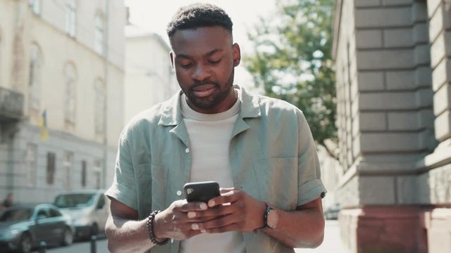 Happy attractive african american man walks down the street Typing on smartphone with interest Enjoying the walk Crowded city on the background sunset sunlight technology