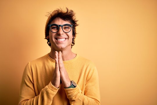 Young handsome man wearing casual t-shirt and glasses over isolated yellow background praying with hands together asking for forgiveness smiling confident.