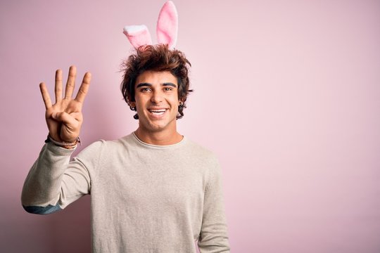 Young handsome man holding easter rabbit ears standing over isolated pink background showing and pointing up with fingers number four while smiling confident and happy.