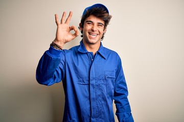 Young mechanic man wearing blue cap and uniform standing over isolated white background smiling positive doing ok sign with hand and fingers. Successful expression.