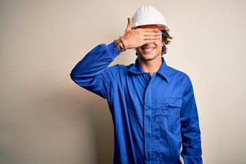 Young constructor man wearing uniform and security helmet over isolated white background smiling and laughing with hand on face covering eyes for surprise. Blind concept.