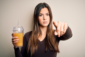 Young beautiful girl drinking glass of healthy orange juice over isolated white background pointing with finger to the camera and to you, hand sign, positive and confident gesture from the front