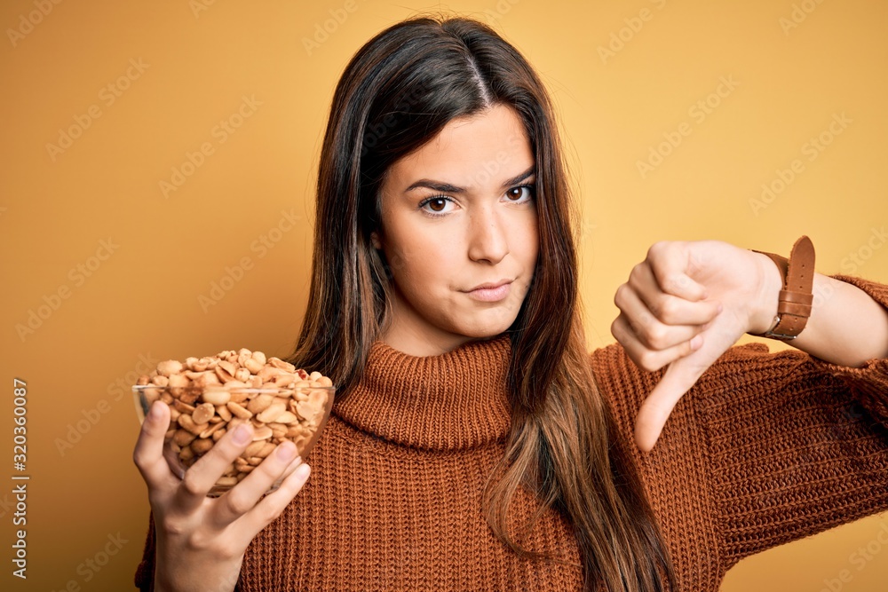 Canvas Prints Young beautiful girl holding bowl of salty peanuts standing over yellow background with angry face, negative sign showing dislike with thumbs down, rejection concept