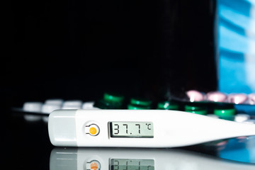 medical jars, thermometer, syringe, gauze mask and pills medicines, medicine on a thermometer 37.7 degrees Celsius on glass black background close up. copy space