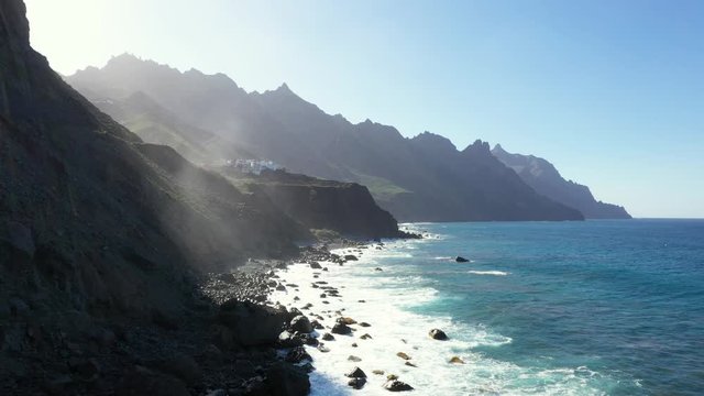 Beautiful coastline surrounded with mighty peaky cliffs and waves hitting the black rocky coast with lens flare on a bright day in Tenerife , Spain canary islands. Drone shot.