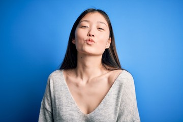 Obraz premium Young beautiful asian woman wearing casual sweater standing over blue isolated background looking at the camera blowing a kiss on air being lovely and sexy. Love expression.
