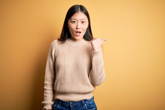Young beautiful asian woman wearing casual sweater over yellow isolated background Surprised pointing with hand finger to the side, open mouth amazed expression.