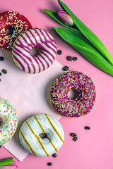 Donuts and coffee on pink background. A breakfast of a cup of coffee and sweet donuts with pink and white icing on a pink background next to the flowers.Top view, copy space, Vertical background