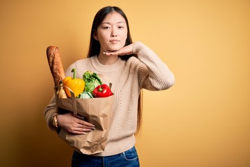 Young asian woman holding paper bag of fresh healthy groceries over yellow isolated background cutting throat with hand as knife, threaten aggression with furious violence