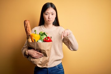 Young asian woman holding paper bag of fresh healthy groceries over yellow isolated background Pointing down looking sad and upset, indicating direction with fingers, unhappy and depressed.