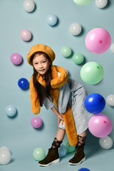 Fototapeta na wymiar Little asian schoolgirl in shirt dress, brown beret and scarf, boots. She smiling, posing with balloons on blue background.