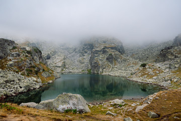 View of a beautiful high mountain lake with calm water surrounded by foggy rocky shore. The Scary Lake, Rila National Park, Bulgaria. 