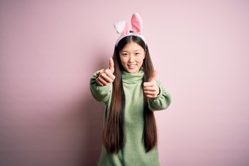 Young asian woman wearing cute easter bunny ears over pink background approving doing positive gesture with hand, thumbs up smiling and happy for success. Winner gesture.