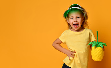 Little smiling blond girl in yellow t-shirt and hat holding fresh fruit juice in pineapple shaped bottle with straw and posing