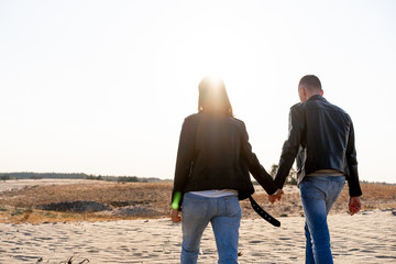 Obraz na płótnie Canvas Young European couple dressed leather jacket and blue jeans walking desert holding hands view from the back.