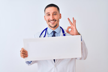 Young doctor man holding blank advertising banner over isolated background doing ok sign with fingers, excellent symbol