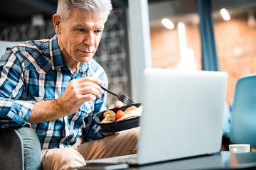 Mature man holding food and looking at laptop screen