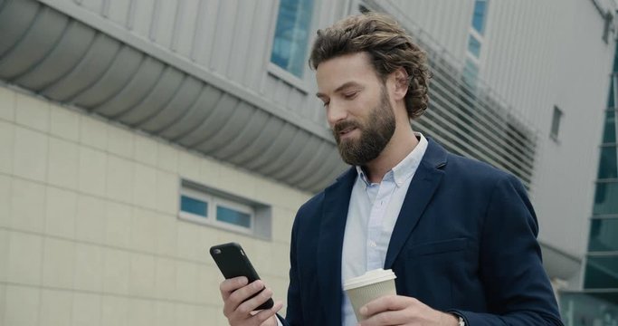 Bearded Businessman is walking near Modern Office Building while Coffe Break. He’s texting message and drinking Coffee to go. Stylish man is wearing smart casual Suit. Men. Apps. Social network.