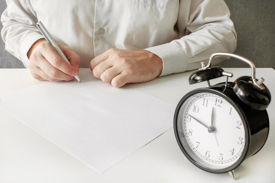 A man puts a signature at the bottom of a blank sheet of document. The contract of sale or power of attorney or testament. Alarm clock close-up with the passing time in the foreground.