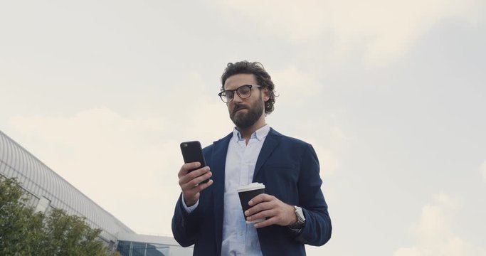 Young handsome scientist in glasses walking near Modern Laboratory. Stylish Man drinking Coffee to go and using his Smartphone while the Walk. Wearing stylish Suit. Professor. Young Teachers. People.