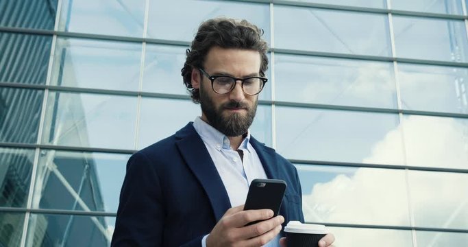 Young confident Professor in Glasses looking to the Camera confidently. Portrait of young Caucasian Man with stylish hair and Look. Using his Smartphone. Social Network. Work outside. Success. People.