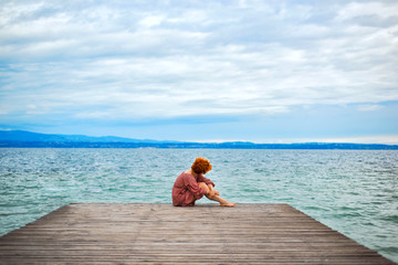 Back of faceless red haired woman in red dress sitting on wooden pier on Garda lake in cloudy day. Italian vacation. Garda lake great tourist destination and attraction in North Italy.