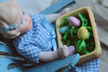 Fototapeta premium A little porcelain baby doll in blue checkered dress holds an Easter basket with colored eggs for a favorite Christian holiday.